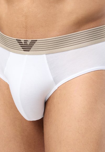 Κοντινή λήψη ενός ατόμου που φοράει λευκά εσώρουχα με μπεζ μέση που έχει ριγέ μοτίβο και μαύρο γεωμετρικό λογότυπο.
