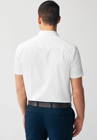 Camisa blanca de manga corta con cuello clásico, vista desde la parte de atrás, combinada con pantalones azul marino y un cinturón marrón. Textura de tela suave.