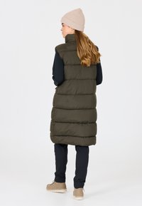 Dunkelgrüne Steppweste, knielang, mit geripptem Kragen, kombiniert mit schwarzen Hosen und beigen Stiefeln, die ein einfaches, funktionales Design einfangen.