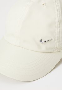Balta cepure, izgatavota no gludas auduma, ar izliekto zīmi. Priekšējā kreisajā pusē atrodas metālisks Nike logotips.