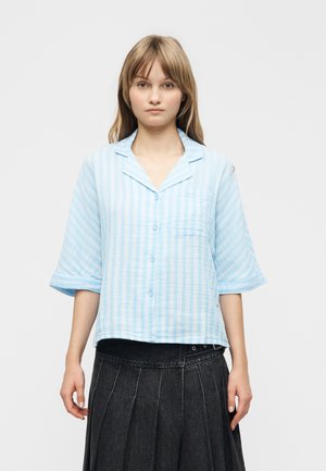 Joven con cabello lacio castaño claro, vestida con una camisa de rayas azul claro abotonada y falda oscura plisada, posando frente a un fondo blanco.