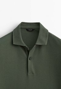 Polo-shirt vert foncé en tissu texturé à mailles en relief. Doté d'une patte de boutonnage et d'un col, présenté à plat sur un fond blanc.