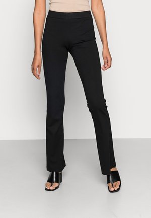 Pantalon noir ajusté avec une taille haute, fabriqué à partir d'un tissu stretch lisse. Associé à des sandales à talons ouverts noires.