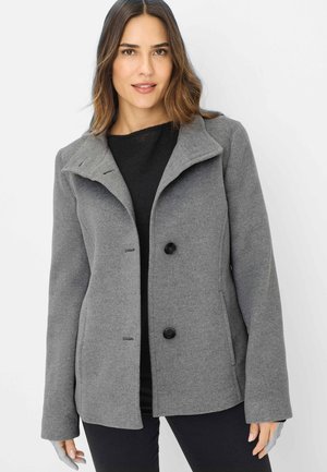 Frau mit schulterlangem braunem Haar, die einen grauen, geknöpften Mantel über einem schwarzen Oberteil trägt und vor einem schlichten hellen Hintergrund steht.