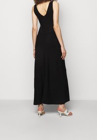 Svart maxiklänning med figursydd bodice och V-back. Materialet verkar ha en textur. Bärs med vita klacksandaler. Enkel design, inga mönster.