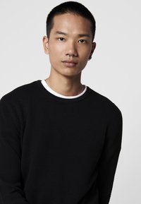 Maglione nero texturizzato con scollo rotondo, indossato sopra una camicia bianca. Primo piano di una persona con capelli corti e un piercing al setto.