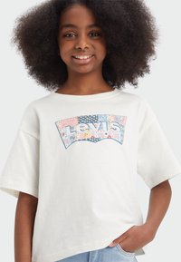 Balts lielgabarīta T-krekls, kas izgatavots no kokvilnas un tam ir krāsains Levi's logo mozaīkas stilā. Īsas piedurknes un brīva kakla izgriezuma forma.
