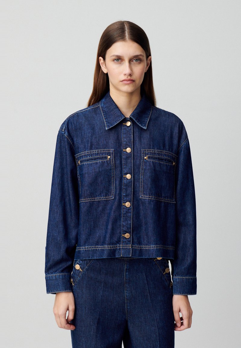 Chaqueta de denim recortada en azul oscuro, con cuello puntiagudo, cierre frontal con botones, dos bolsillos en el pecho y acentos de botones dorados.