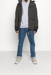 Giacca nera imbottita con cappuccio, chiusura con zip e tasche, indossata sopra un felpa leggera grigia, abbinata a jeans blu e sneakers beige.