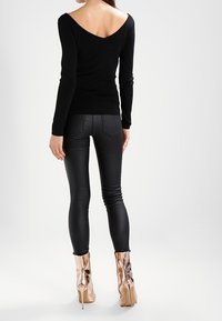 Zwarte langemouwen top met een wijde V-hals en een strakke pasvorm, gecombineerd met glanzende zwarte leggings. Het model draagt metallic hoge hakken.