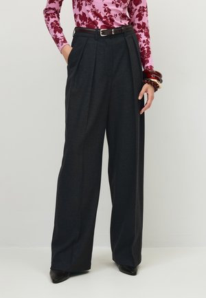 Vrouw met hoge taille, wijde zwarte broek, bloemige top met lange mouwen, zwarte riem, zwarte puntige schoenen en rode en witte armbanden.