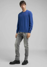 Maglione girocollo blu, jeans grigi aderenti e stivaletti neri alla caviglia. Tessuto liscio con un dettaglio logo sottile sul petto.