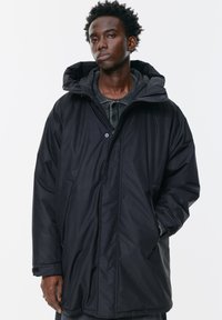 Tânăr îmbrăcat într-o parka de iarnă oversized neagră, cu glugă, stând în fața unui fundal alb simplu, cu o mână în buzunar.