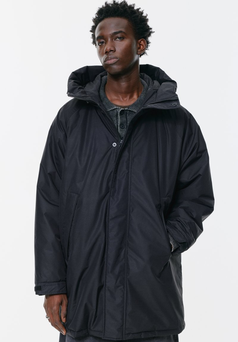 Tânăr îmbrăcat într-o parka de iarnă oversized neagră, cu glugă, stând în fața unui fundal alb simplu, cu o mână în buzunar.