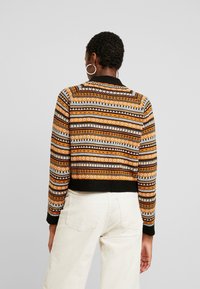 Neulepusero, jossa on monivärinen geometrinen kuvio oranssissa, valkoisessa ja mustassa, lyhyt malli sekä ribbireunat hihoissa ja helmassa.