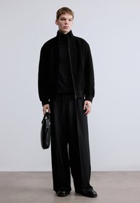 Μαύρο μπουφάν bomber από σουέντ με ψηλό γιακά, συνδυασμένο με μαύρο ζιβάγκο και μαύρο παντελόνι φαρδιάς γραμμής. Κρατάει μαύρη τσάντα χειρός.