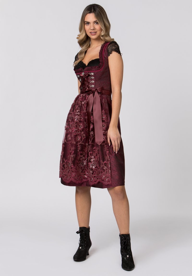 Burgundy dirndl dress with lace cap sleeves, fitted bodice, and embroidered detailing. Satin ribbon waist tie, knee-length skirt, and skirt hem accents.
