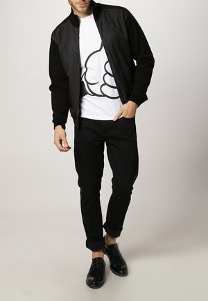 Mężczyzna ubrany w czarną kurtkę, białą koszulkę z dużym czarnym konturem ręki, czarne spodnie i czarne eleganckie buty na jednolitym tle.