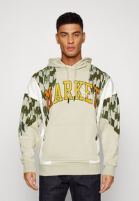 Felpa con cappuccio in beige chiaro, con testo arancione "MARKET", maniche in camouflage verde e dettagli bianchi, tasca frontale e stringhe di regolazione.