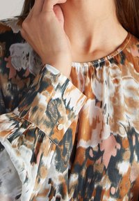 Main d'une femme touchant son cou, portant un chemisier à motif abstrait multicolore aux tons orange, noir, blanc et beige.