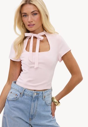 Jeune femme aux longs cheveux blonds portant un haut rose pâle avec une encolure en trou de serrure et un nœud, un jean bleu clair taille haute, et un bracelet en or.