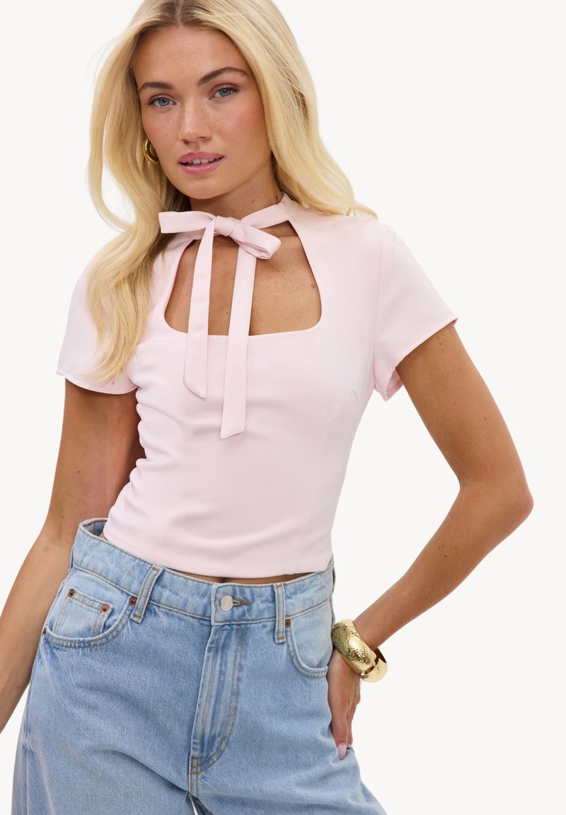 Jeune femme aux longs cheveux blonds portant un haut rose pâle avec une encolure en trou de serrure et un nœud, un jean bleu clair taille haute, et un bracelet en or.