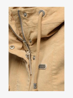 Gros plan sur la fermeture éclair d'une veste beige, un cordon tressé, des boutons-pression en métal et une petite plaque de marque rectangulaire sur le tissu.