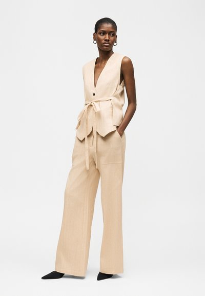 Femme debout, les mains dans les poches, portant un gilet sans manches beige ceinturé et un pantalon large assorti, des chaussures noires à bouts pointus et des boucles d'oreilles créoles.