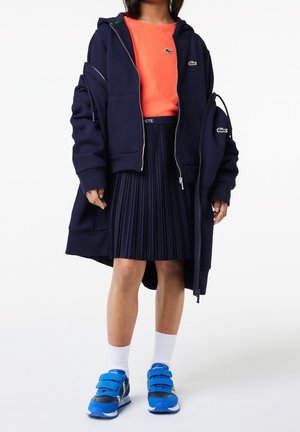 Felpa con cappuccio blu navy con zip, indossata sopra una t-shirt arancione, gonna plissettata blu navy, calzini bianchi e sneakers blu con chiusura a velcro e dettagli neri.