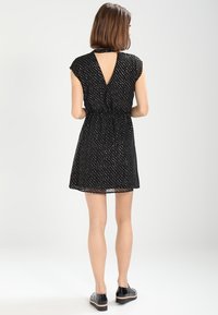 Femme avec les cheveux bruns courts portant une robe noire à manches courtes avec des pois blancs et une découpe en V dans le dos, associée à des chaussures à plateforme noires.