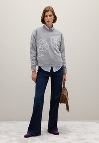 Vrouw in een grijze trui met bloemmotief over een gestreept shirt, donker flare-jeans, paarse schoenen en met een bruine handtas met franjes.