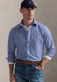 Blauw-witte geruite shirt met lange mouwen, gecombineerd met een wit T-shirt, een bruine gevlochten leren riem en een marineblauwe pet.
