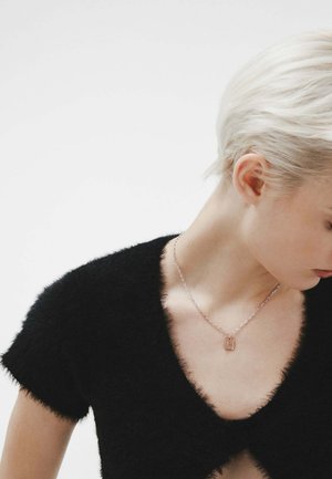 Mujer con cabello rubio corto que lleva un top negro con cuello en V de textura suave y un delicado collar de cadena con un pequeño colgante rectangular.