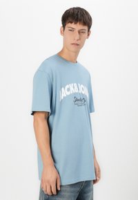 Ung mand i lyseblå oversized T-shirt med "JACK&JONES" logo og blå jeans, der står foran en ensfarvet hvid baggrund og kigger til højre.