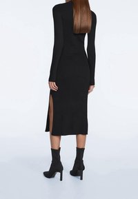 Abito nero a coste con maniche lunghe, dotato di spacchi laterali all'orlo. Abbinato a stivaletti neri e una silhouette elegante.