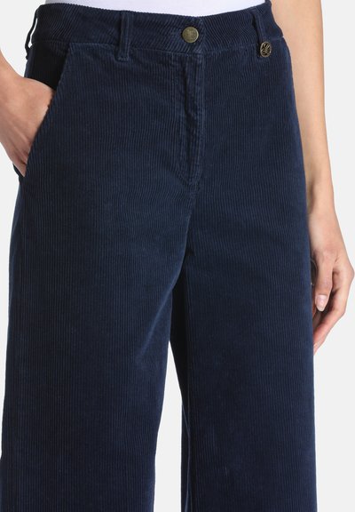 Pantalon en velours côtelé bleu marine avec poches avant, passants de ceinture et fermeture par bouton, porté par une personne avec une main dans la poche.