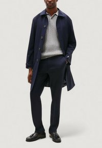 Cappotto trench navy su un maglione grigio chiaro a trama cable-knit, abbinato a pantaloni scuri e mocassini neri. Focus sul design elegante e sul taglio su misura.