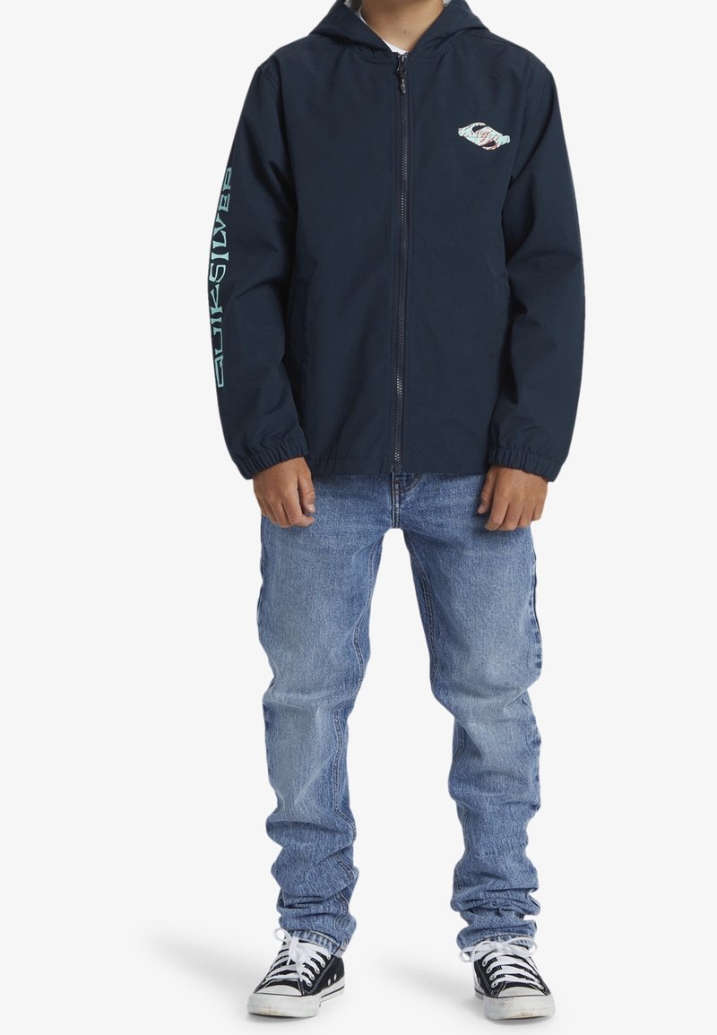 Garçon portant un sweat à capuche bleu marine zippé avec un logo sur la poitrine et la manche, un jean bleu clair et des baskets noires et blanches, debout devant un fond blanc.