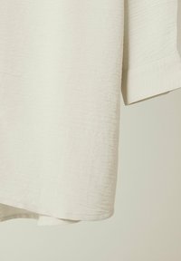 Light beige short-sleeve shirt made of textured fabric, featuring a rounded hem and clean stitching at the sleeves and bottom edge.