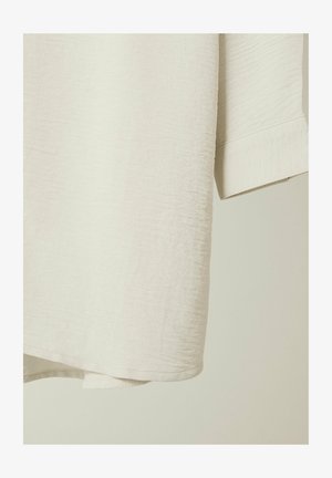 Lys beige kortærmet skjorte lavet af struktureret stof, med en rund nederkant og ren syning ved ærmerne og nederste kant.