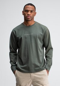 Man in olijfgroene lange mouwen shirt met de tekst "BUTCHER OF BLUE" en beige cargobroek, staande met handen in zijn zakken tegen een witte achtergrond.