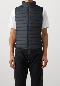 Pufferväst i mörkgrått, med ståkrage, dragkedja och horisontell quilt-design; bärs över en vit skjorta.