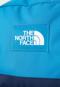 Un tissu bleu avec une surface texturée présente un écusson rectangulaire blanc portant l'inscription "THE NORTH FACE" et un accent en demi-cercle.