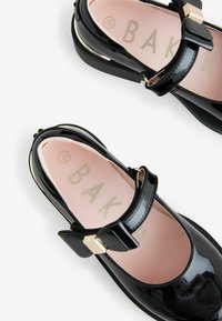 Zwarte lakleren schoenen met een ronde neus, enkelbandje en gouden accenten. Roze binnenzool met de opdruk "BAK." Ontworpen voor comfort.