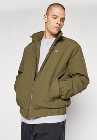 Chaqueta de color verde oliva con cremallera y cuello alzado, puños y cintura elásticos; presenta un pequeño logo en el pecho. Hecha de material ligero.