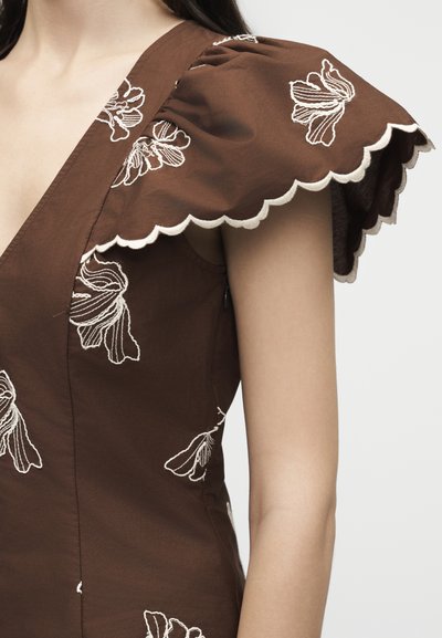 Καφέ φόρεμα με φουσκωτά μανίκια με κυματιστές άκρες και λευκή κεντημένη ανθοδέσμη πάνω στο ύφασμα, φορεμένο από άτομο με ακάλυπτα χέρια.