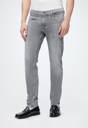 Mies pukeutunut vaaleanharmaisiin slim fit -farkkuihin, mustiin nahkalaiskaoloihin ja valkoisiin sukkiin yksiväristä taustaa vasten.