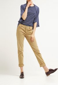 Hnědé bavlněné kalhoty s ohrnutými nohavicemi, které jsou kombinovány s námořnickým modrým a bílým pruhovaným tričkem a volným modrým kardiganem. Hnědé šněrovací boty dotvářejí celý vzhled.