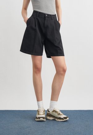 Persona que lleva pantalones cortos plisados negros, camiseta sin mangas gris, calcetines blancos hasta el tobillo y zapatillas beige y marrones de pie sobre una alfombra azul.