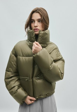 Femme portant une doudoune vert olive à col montant, tenant la fermeture éclair près du menton, debout sur un fond clair uni.